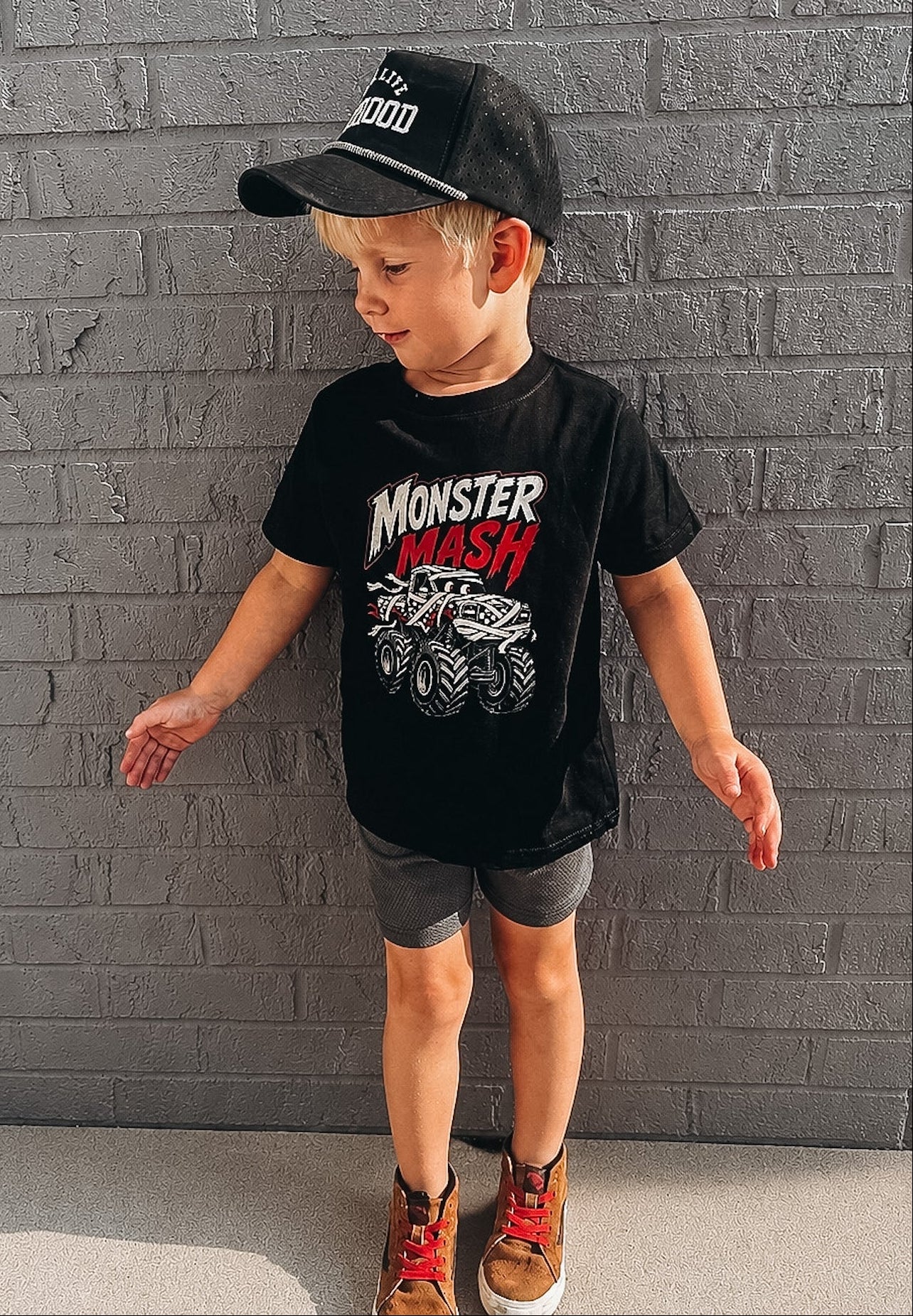 A young boy wearing a black t-shirt with a 'Monster Mash' monster truck graphic, gray shorts, and a black baseball cap, standing against a gray brick wall.