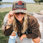 A woman outdoors wearing a brown trucker hat with a 'Mom's Group' patch and a red rose graphic, paired with a brown textured jacket and ripped blue jeans.