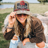 A woman outdoors wearing a brown trucker hat with a 'Mom's Group' patch and a red rose graphic, paired with a brown textured jacket and ripped blue jeans.