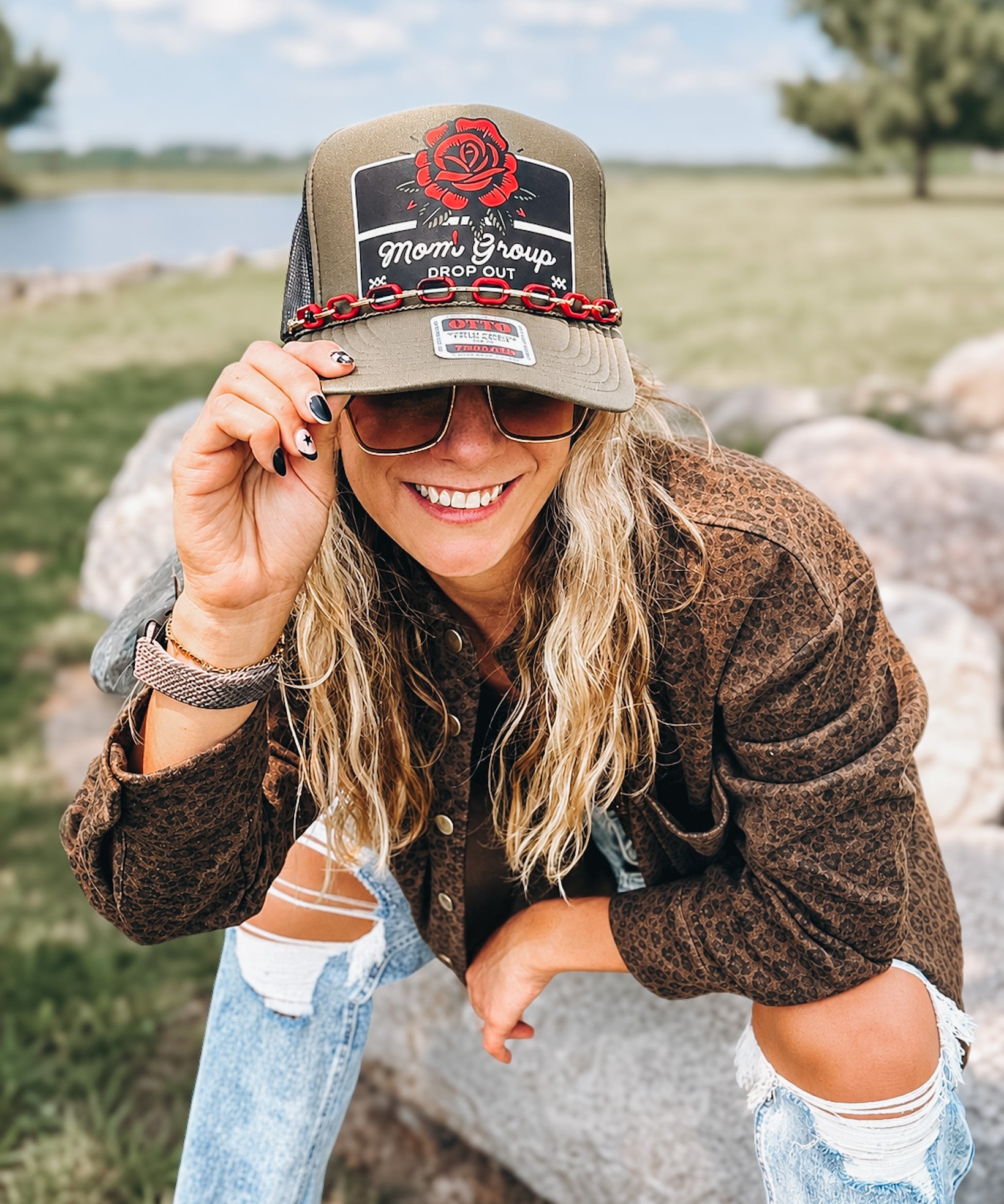 A woman outdoors wearing a brown trucker hat with a 'Mom's Group' patch and a red rose graphic, paired with a brown textured jacket and ripped blue jeans.
