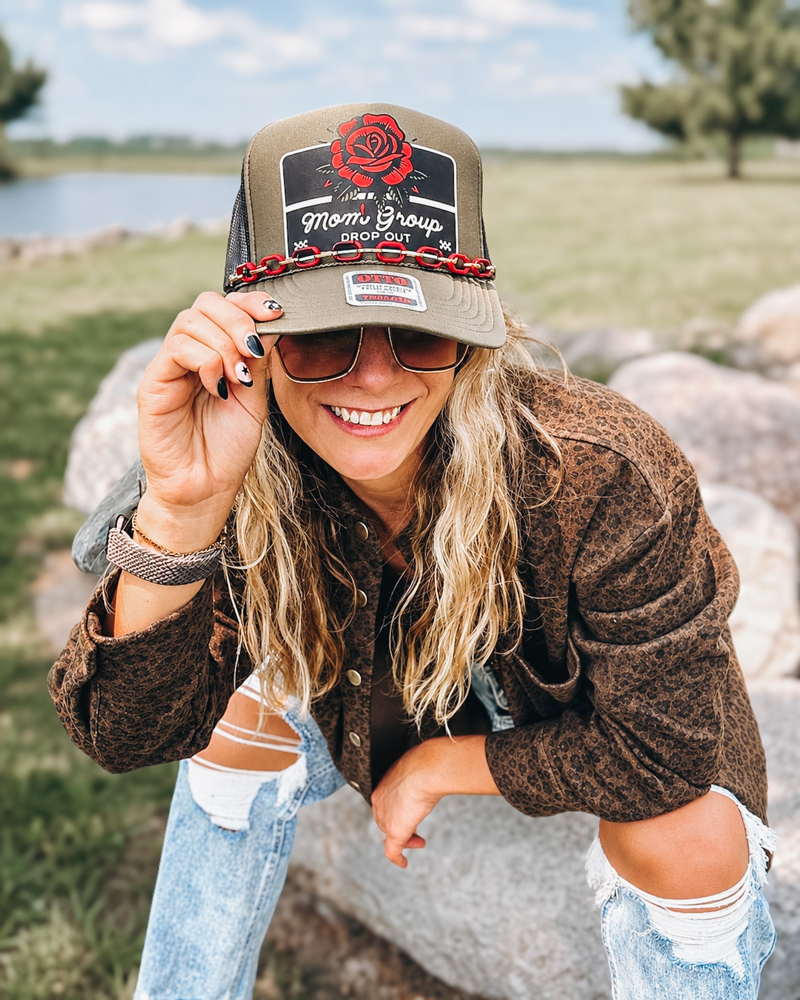 A woman outdoors wearing a brown trucker hat with a 'Mom's Group' patch and a red rose graphic, paired with a brown textured jacket and ripped blue jeans.