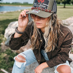 A woman wearing a brown leopard-print jacket, distressed light-wash jeans, and an olive green trucker hat with a red rose graphic and 'Stoner Group' text, sitting outdoors on a rock near water.