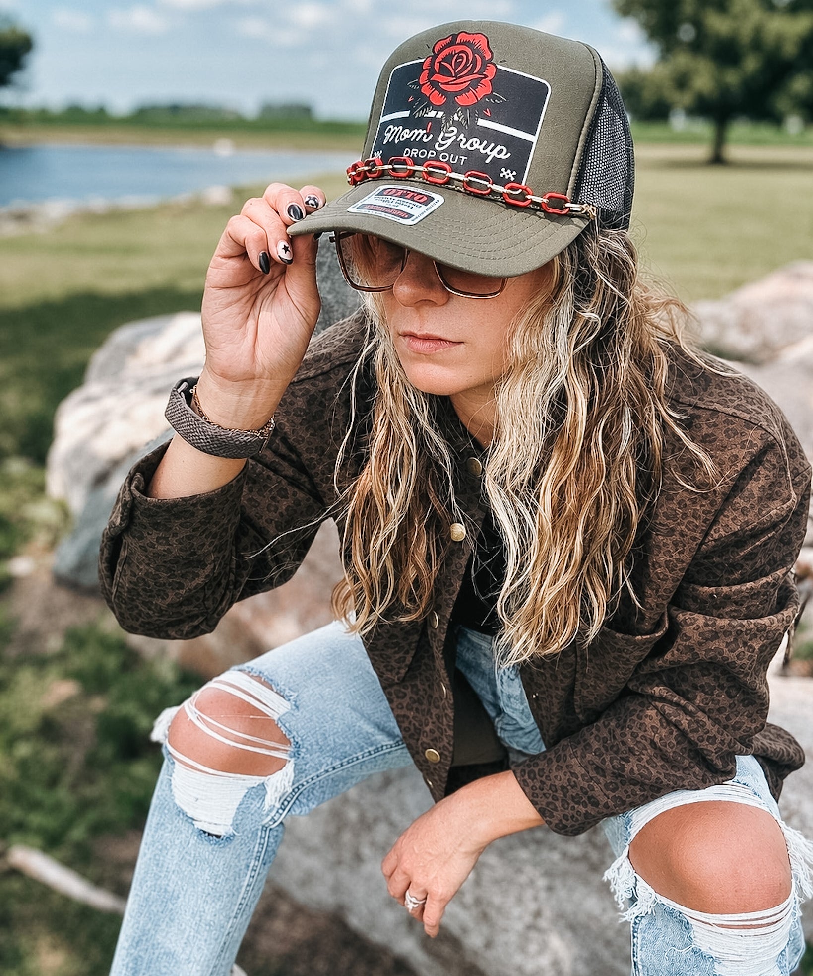 A woman wearing a brown leopard-print jacket, distressed light-wash jeans, and an olive green trucker hat with a red rose graphic and 'Stoner Group' text, sitting outdoors on a rock near water.