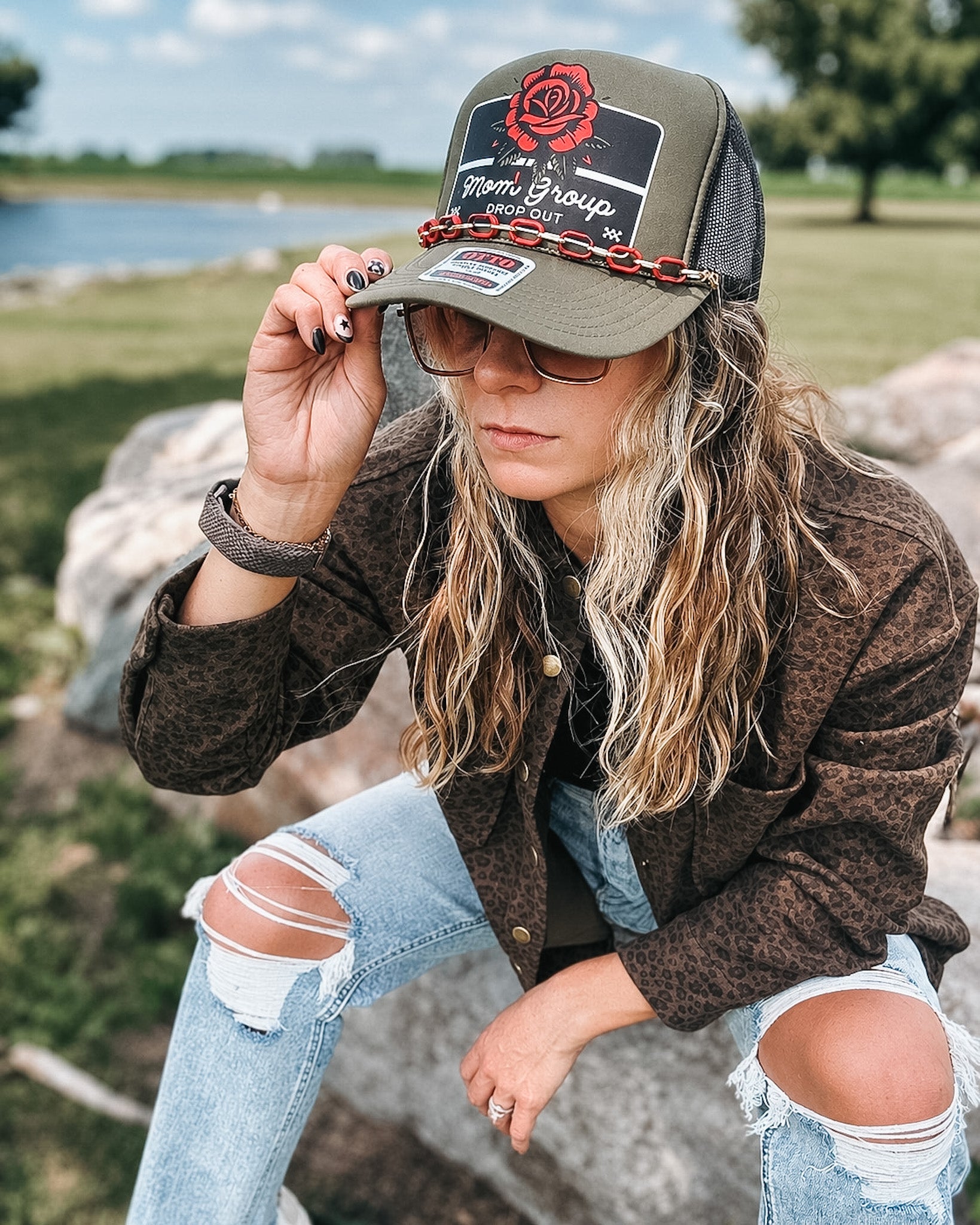 A woman wearing a brown leopard-print jacket, distressed light-wash jeans, and an olive green trucker hat with a red rose graphic and 'Stoner Group' text, sitting outdoors on a rock near water.