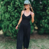 A woman wearing a black sleeveless jumpsuit, black sandals, and a green baseball cap with 'HERS' written on it, standing outdoors on a grassy lawn with a green hedge in the background.