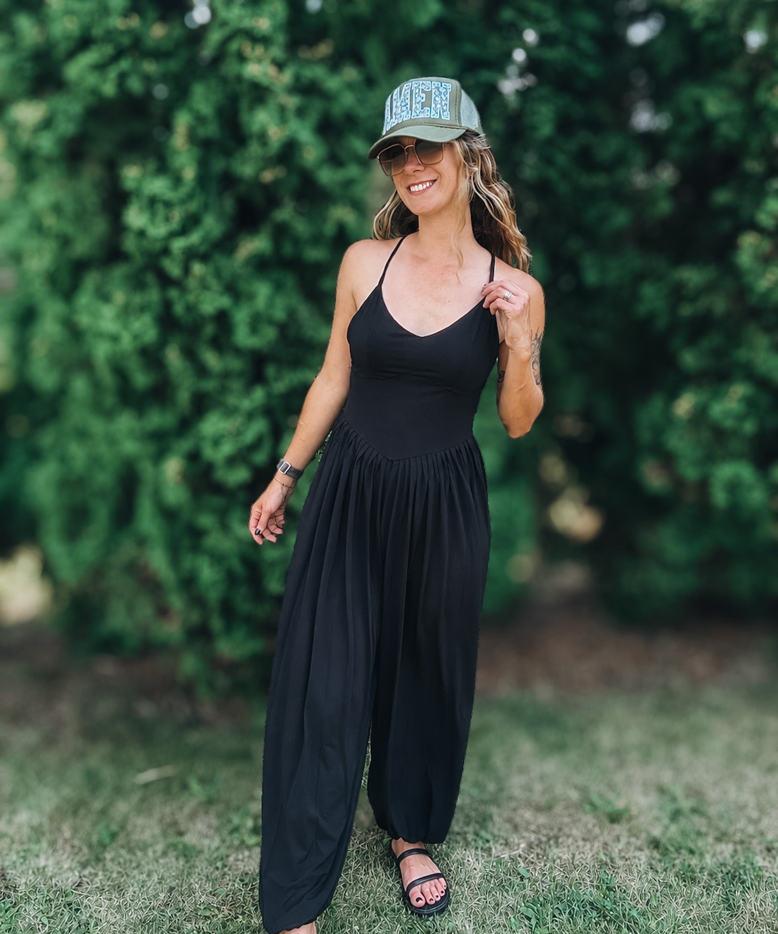 A woman wearing a black sleeveless jumpsuit, black sandals, and a green baseball cap with 'HERS' written on it, standing outdoors on a grassy lawn with a green hedge in the background.