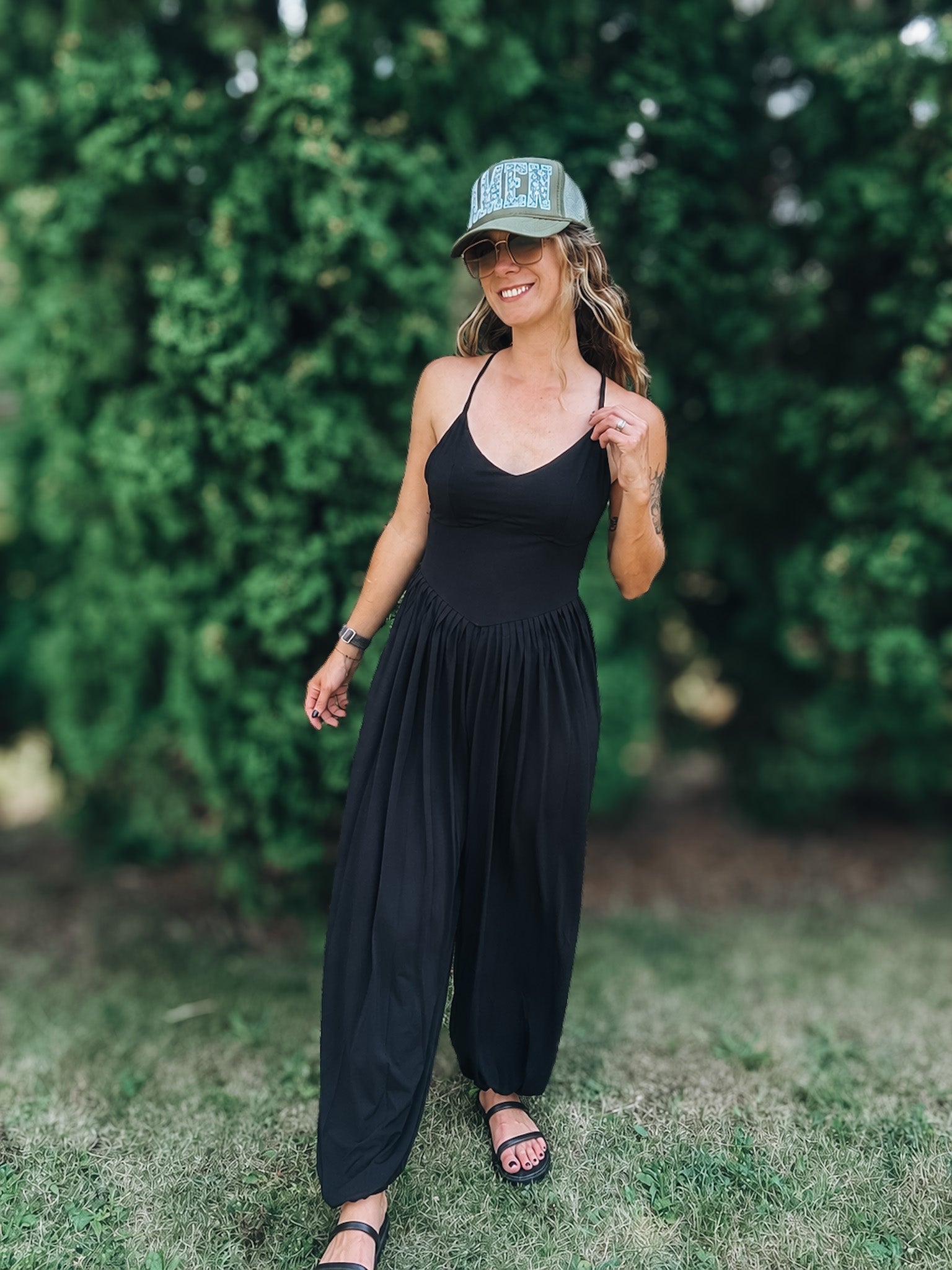 A woman wearing a black sleeveless jumpsuit, black sandals, and a green baseball cap with 'HERS' written on it, standing outdoors on a grassy lawn with a green hedge in the background.