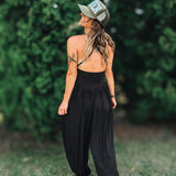 A woman wearing a black backless jumpsuit with wide-leg harem pants, black strappy sandals, and an olive green trucker hat, standing outdoors on grass with a blurred green hedge background.
