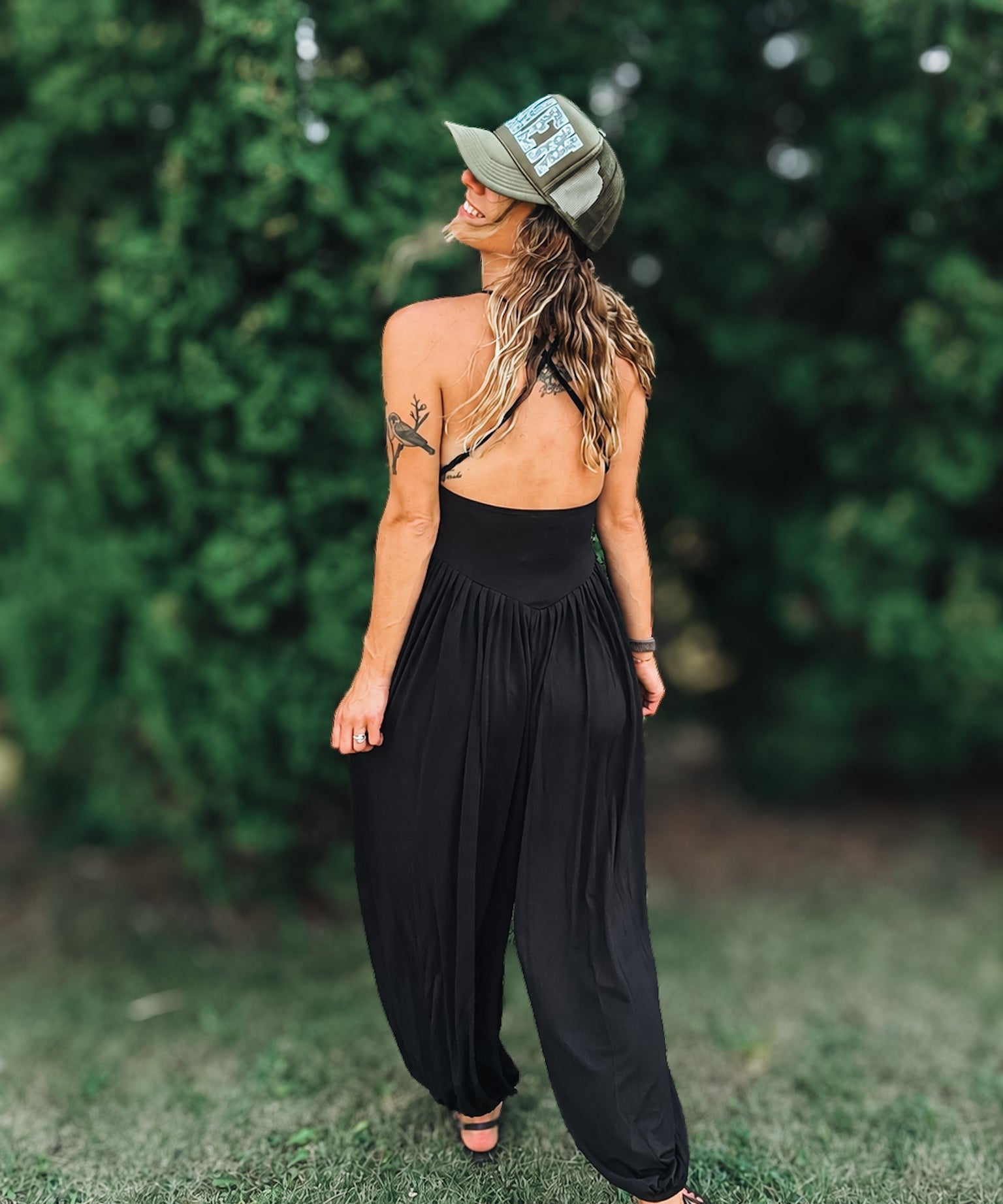 A woman wearing a black backless jumpsuit with wide-leg harem pants, black strappy sandals, and an olive green trucker hat, standing outdoors on grass with a blurred green hedge background.