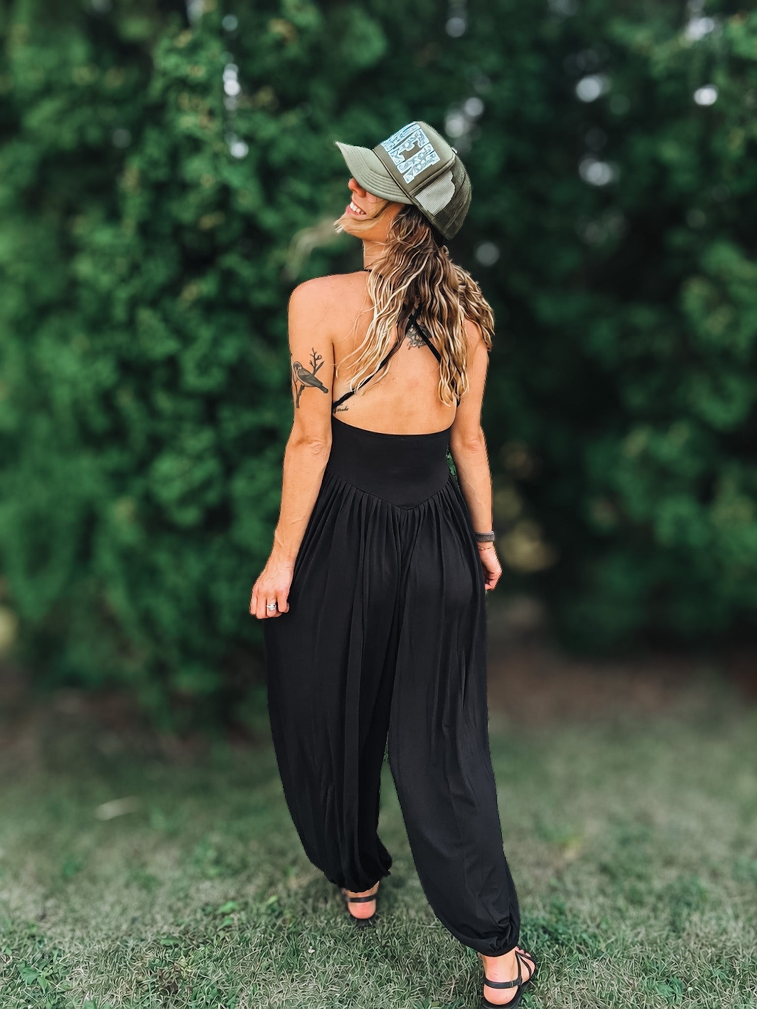 A woman wearing a black backless jumpsuit with wide-leg harem pants, black strappy sandals, and an olive green trucker hat, standing outdoors on grass with a blurred green hedge background.