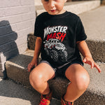 A young boy wearing a black Monster Mash graphic t-shirt, black shorts, and brown sneakers sits on concrete steps outdoors.