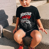 A young boy wearing a black Monster Mash graphic t-shirt, black shorts, and brown sneakers sits on concrete steps outdoors.