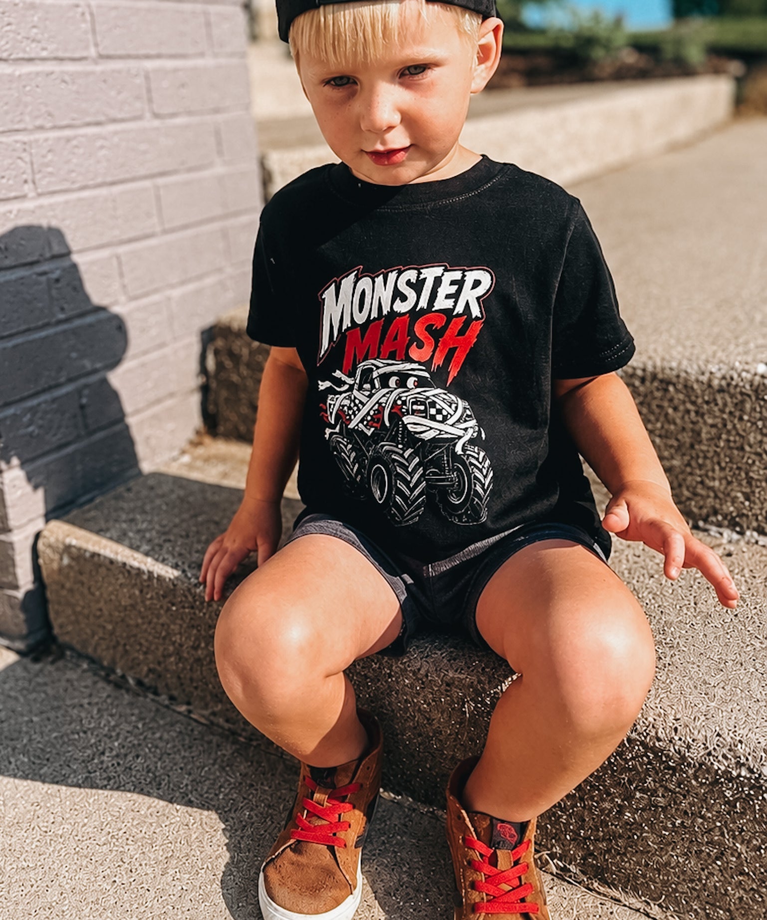 A young boy wearing a black Monster Mash graphic t-shirt, black shorts, and brown sneakers sits on concrete steps outdoors.