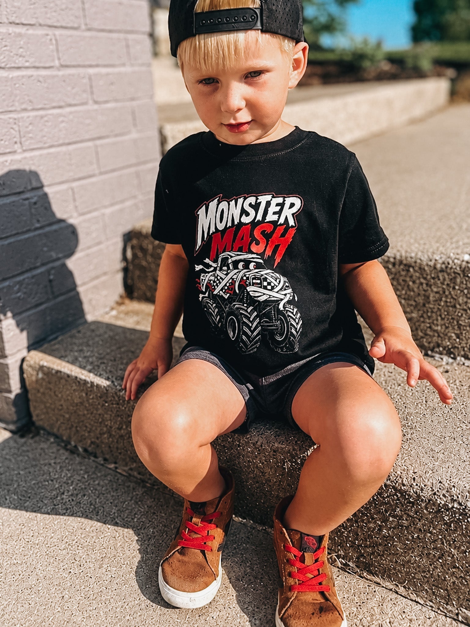 A young boy wearing a black Monster Mash graphic t-shirt, black shorts, and brown sneakers sits on concrete steps outdoors.