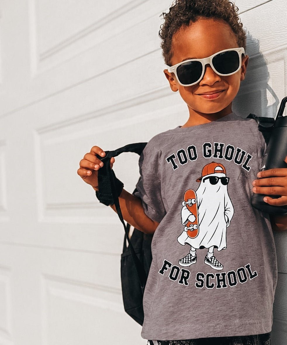 A young child wearing a gray graphic t-shirt with a ghost design and the text 'TOO GHOUL FOR SCHOOL', black patterned shorts, and white-framed sunglasses, posing against a white garage door.