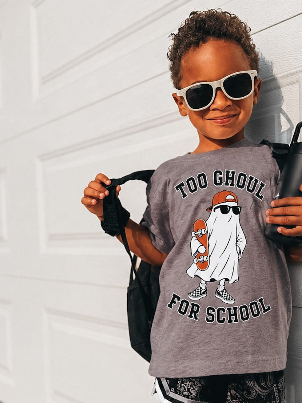 A young child wearing a gray graphic t-shirt with a ghost design and the text 'TOO GHOUL FOR SCHOOL', black patterned shorts, and white-framed sunglasses, posing against a white garage door.