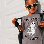 A young child wearing a gray graphic t-shirt with a ghost design and the text 'TOO GHOUL FOR SCHOOL', black patterned shorts, and white-framed sunglasses, posing against a white garage door.