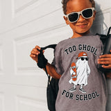 A young child wearing a gray graphic t-shirt with a ghost design and the text 'TOO GHOUL FOR SCHOOL', black patterned shorts, and white-framed sunglasses, posing against a white garage door.