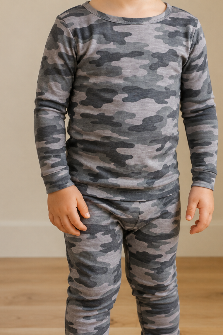 A child wearing a long-sleeve camouflage pajama set in shades of gray and black, standing barefoot on a light wood floor against a beige wall.