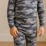 A child wearing a long-sleeve camouflage pajama set in shades of gray and black, standing barefoot on a light wood floor against a beige wall.