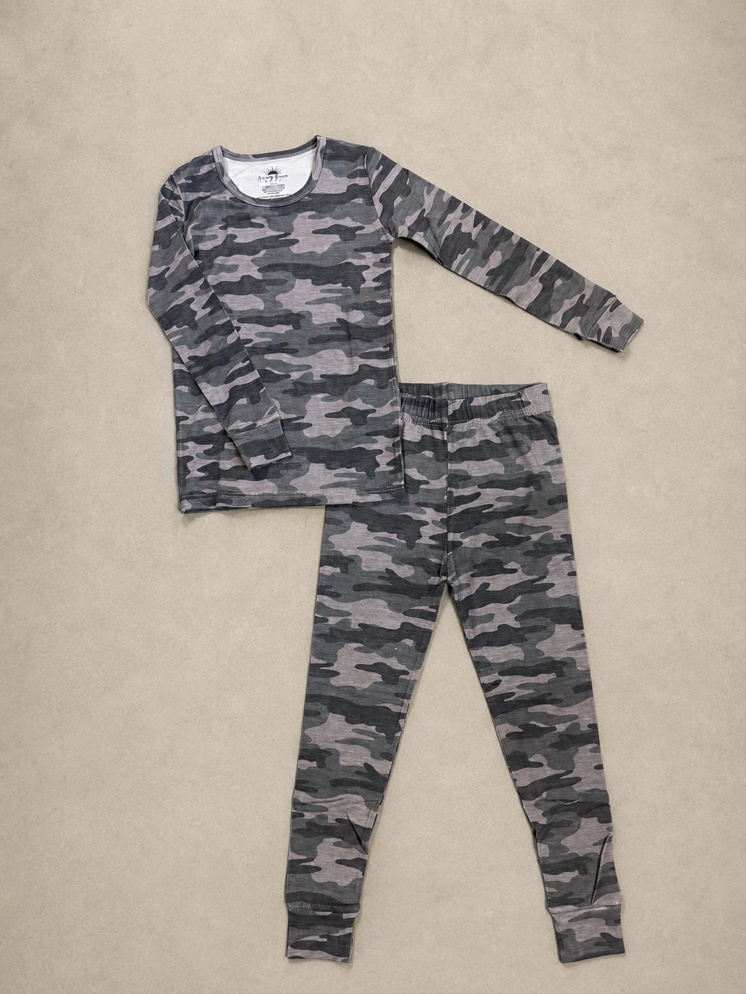 A two-piece children's pajama set featuring a long-sleeve top and matching pants in a gray and black camouflage pattern, laid flat on a beige textured background.