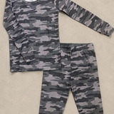 A two-piece children's pajama set featuring a long-sleeve top and matching pants in a gray and black camouflage pattern, laid flat on a beige textured background.