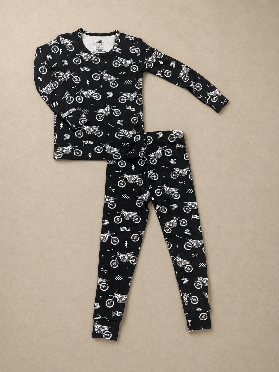 A two-piece children's pajama set featuring a long-sleeve top and matching leggings, both in black with a white allover print of dirt bikes, flags, and road signs, laid flat on a beige background.