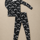 A two-piece children's pajama set featuring a long-sleeve top and matching leggings, both in black with a white allover print of dirt bikes, flags, and road signs, laid flat on a beige background.