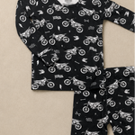 A two-piece children's pajama set featuring a long-sleeve top and matching leggings, both in black with a white allover print of dirt bikes, flags, and road signs, laid flat on a beige background.