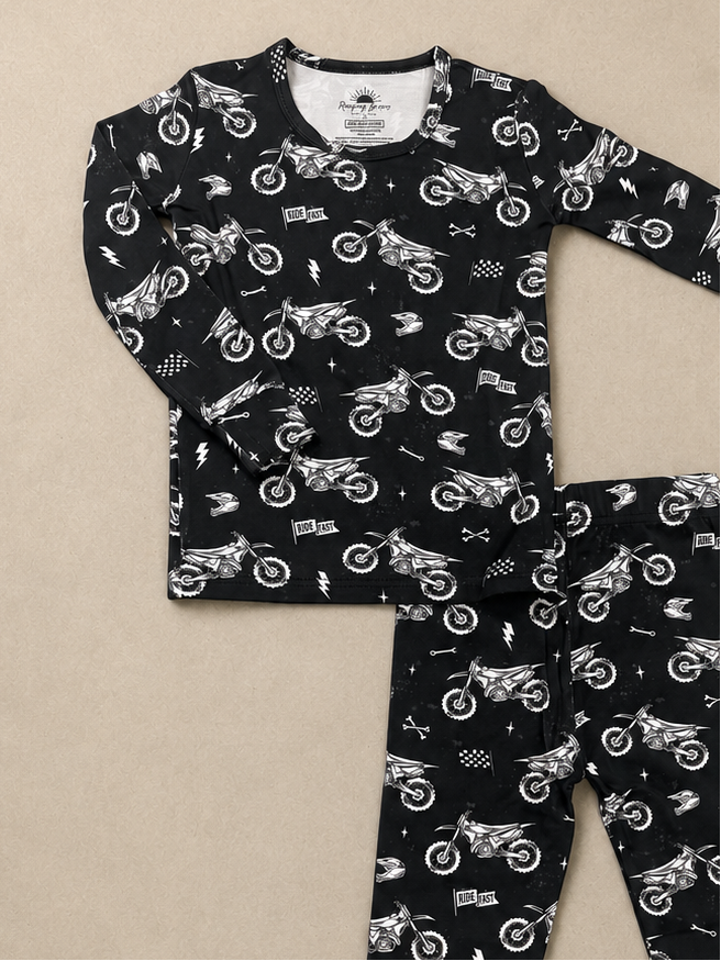 A two-piece children's pajama set featuring a long-sleeve top and matching leggings, both in black with a white allover print of dirt bikes, flags, and road signs, laid flat on a beige background.
