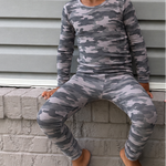 Two young children sitting on a gray brick ledge against a gray siding wall, wearing matching long-sleeve pajamas. The child on the left wears a gray camouflage pattern, and the child on the right wears a dark navy pattern with white circular motifs.
