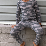 Two young children sitting on a gray brick ledge against a gray siding wall, wearing matching long-sleeve pajamas. The child on the left wears a gray camouflage pattern, and the child on the right wears a dark navy pattern with white circular motifs.