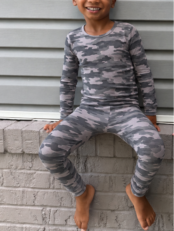 Two young children sitting on a gray brick ledge against a gray siding wall, wearing matching long-sleeve pajamas. The child on the left wears a gray camouflage pattern, and the child on the right wears a dark navy pattern with white circular motifs.