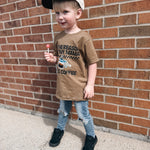 Child wearing a brown t-shirt with text and a cap, standing against a brick wall.