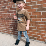 Child wearing a brown t-shirt with text and a cap, standing against a brick wall.