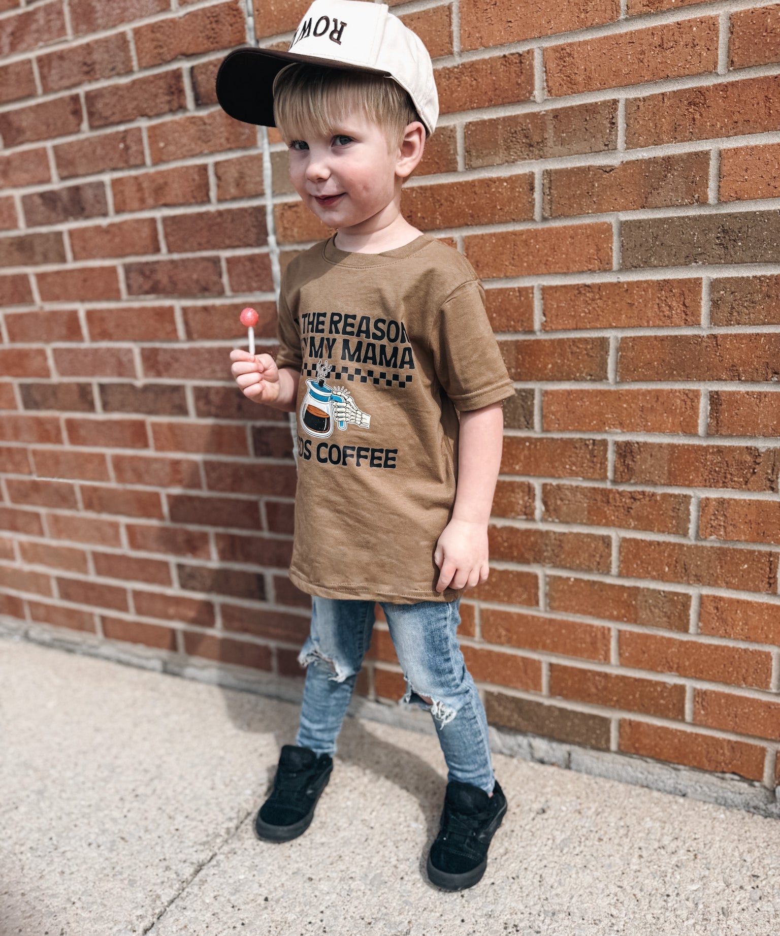 Child wearing a brown t-shirt with text and a cap, standing against a brick wall.