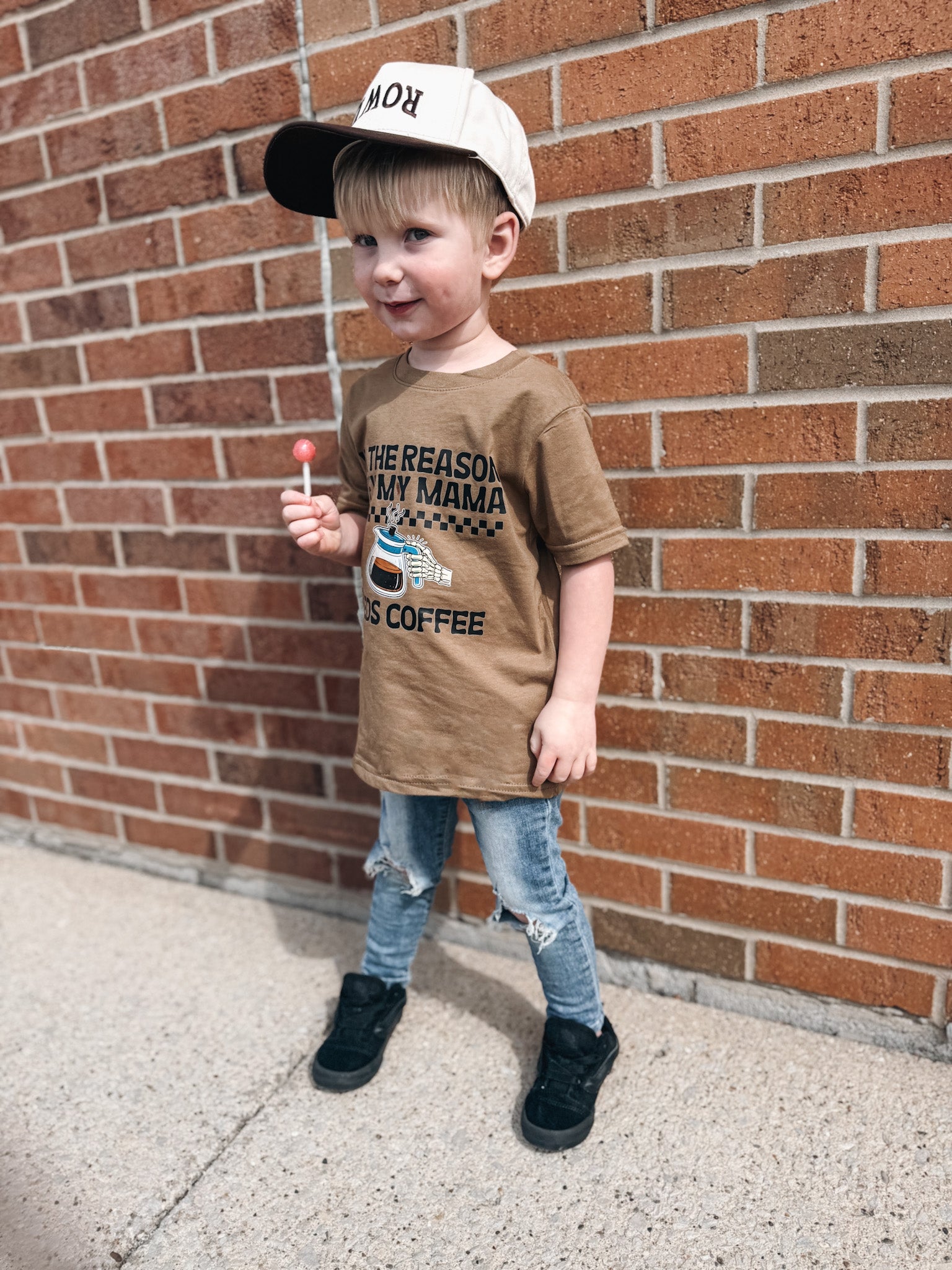 Child wearing a brown t-shirt with text and a cap, standing against a brick wall.