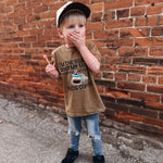 Child wearing a brown t-shirt with a graphic design, standing against a brick wall.