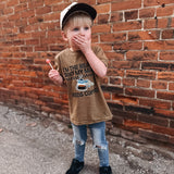 Child wearing a brown t-shirt with a graphic design, standing against a brick wall.