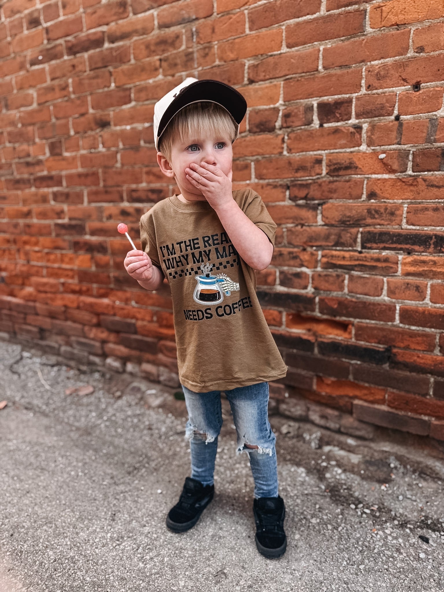 Child wearing a brown t-shirt with a graphic design, standing against a brick wall.