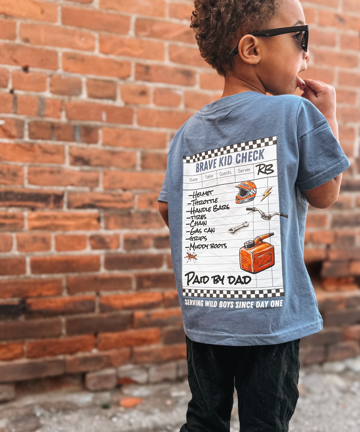Child wearing a blue t-shirt with a graphic design, standing against a brick wall.