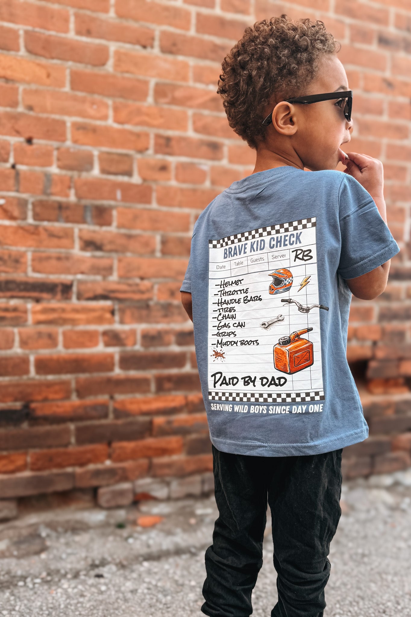 Child wearing a blue t-shirt with a graphic design, standing against a brick wall.