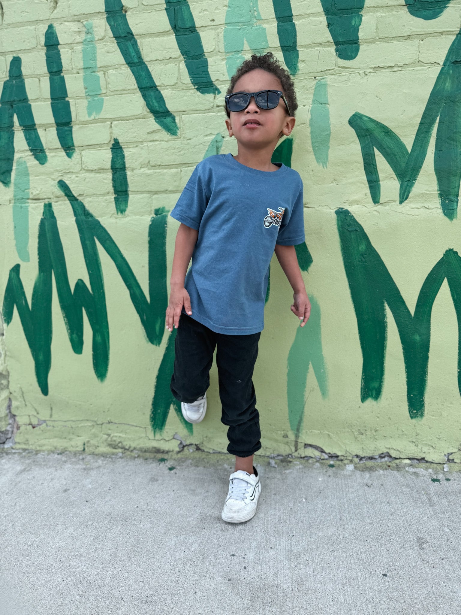Child wearing sunglasses and a blue shirt standing in front of a green mural.