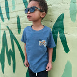 Child wearing sunglasses and a blue t-shirt with a graphic design, standing against a green and beige wall.