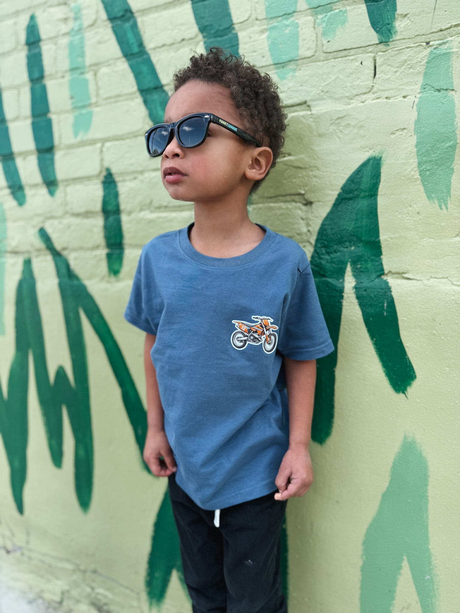 Child wearing sunglasses and a blue t-shirt with a graphic design, standing against a green and beige wall.