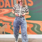 Person wearing a patterned shirt and acid-wash jeans standing in front of a colorful mural.