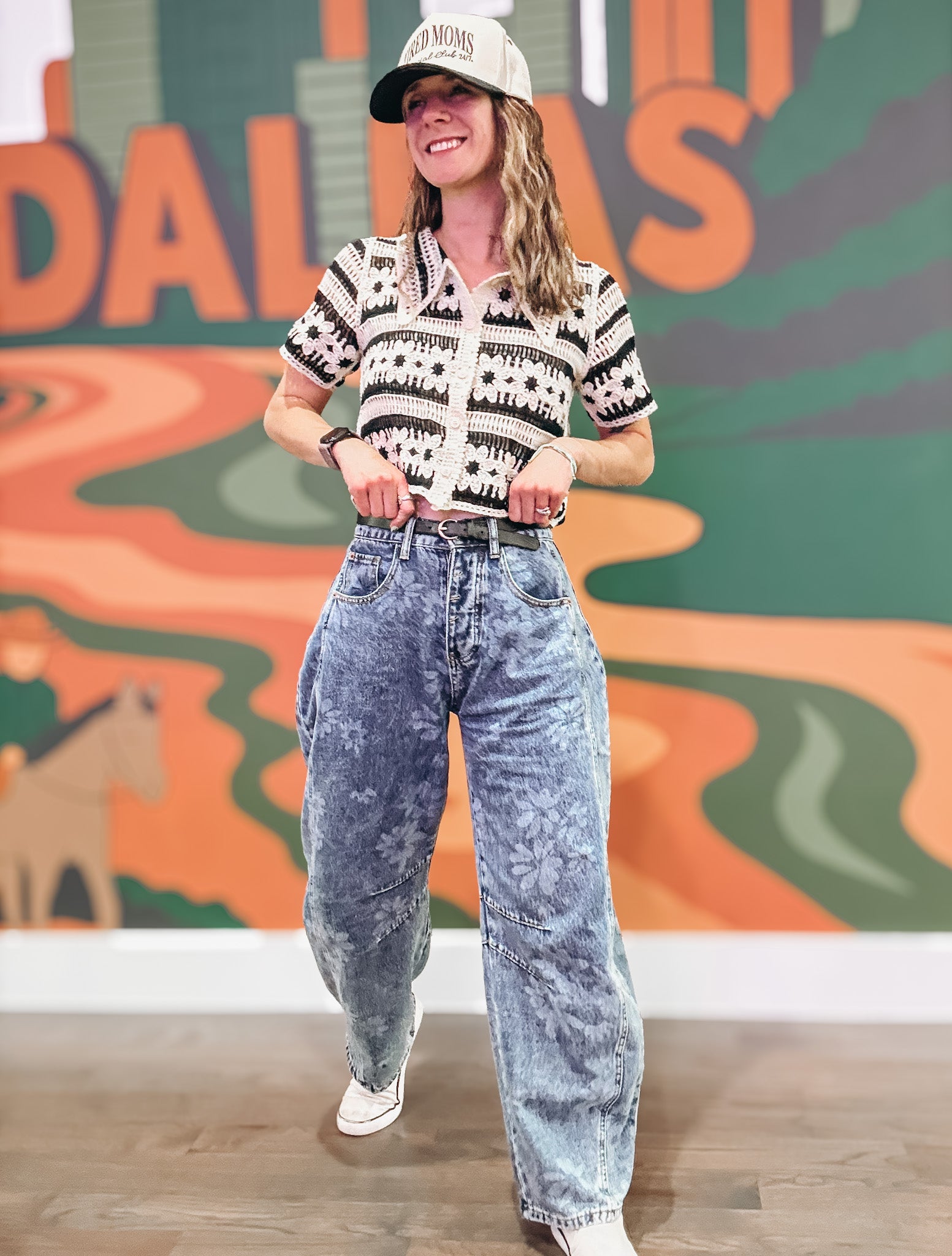 Person wearing a patterned shirt and acid-wash jeans standing in front of a colorful mural.