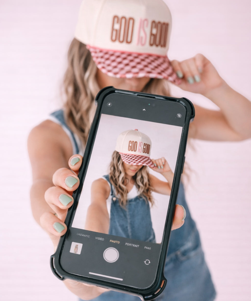 Person wearing a 'GOD IS GOOD' cap, holding a phone with a similar cap displayed on the screen.