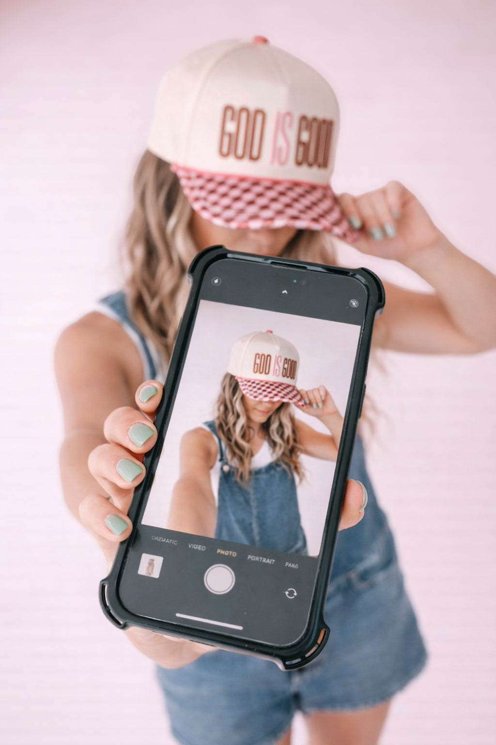 Person wearing a 'GOD IS GOOD' cap, holding a phone with a similar cap displayed on the screen.