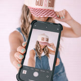 Person wearing a 'GOD IS GOOD' cap, holding a phone with a similar cap displayed on the screen.